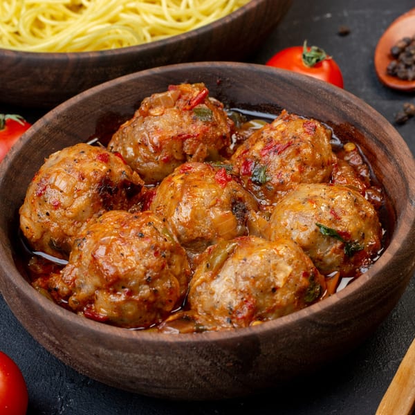 Close-up of meatballs in a rich tomato sauce, with spaghetti and tomatoes in the background.
