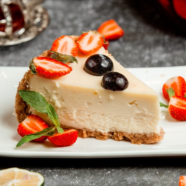 A slice of cheesecake with a graham cracker crust, topped with fresh strawberries, blueberries, and mint.