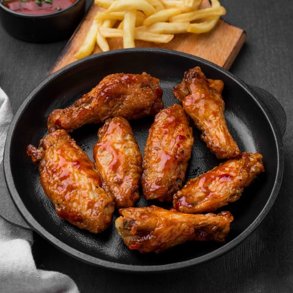 Seven glazed chicken wings in a black skillet with fries and ketchup in the background.