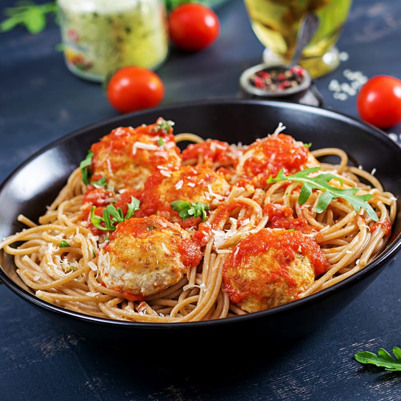 Spaghetti with meatballs, tomato sauce, parmesan, and arugula in a black bowl.